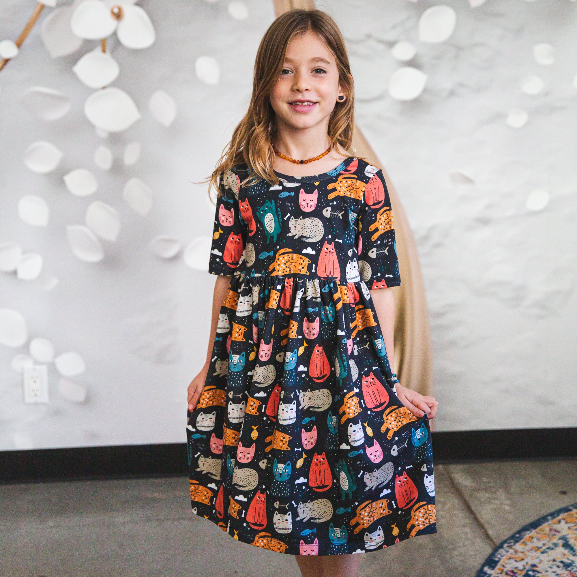 Young girl wearing a dress with cat pattern against a white wall.